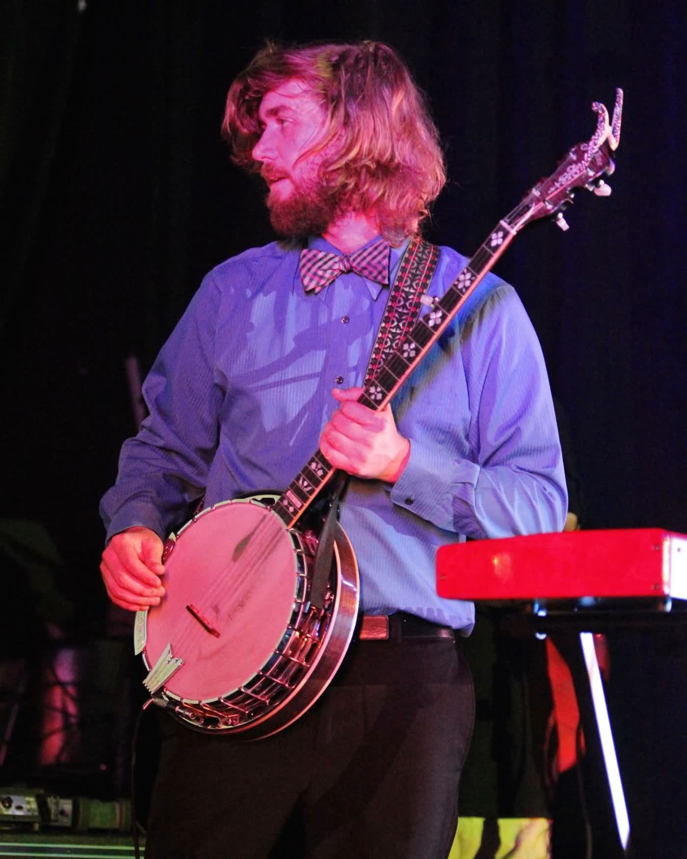 Color photo of Tyler Rice standing with his banjo in a blue shirt and gingham bow tie