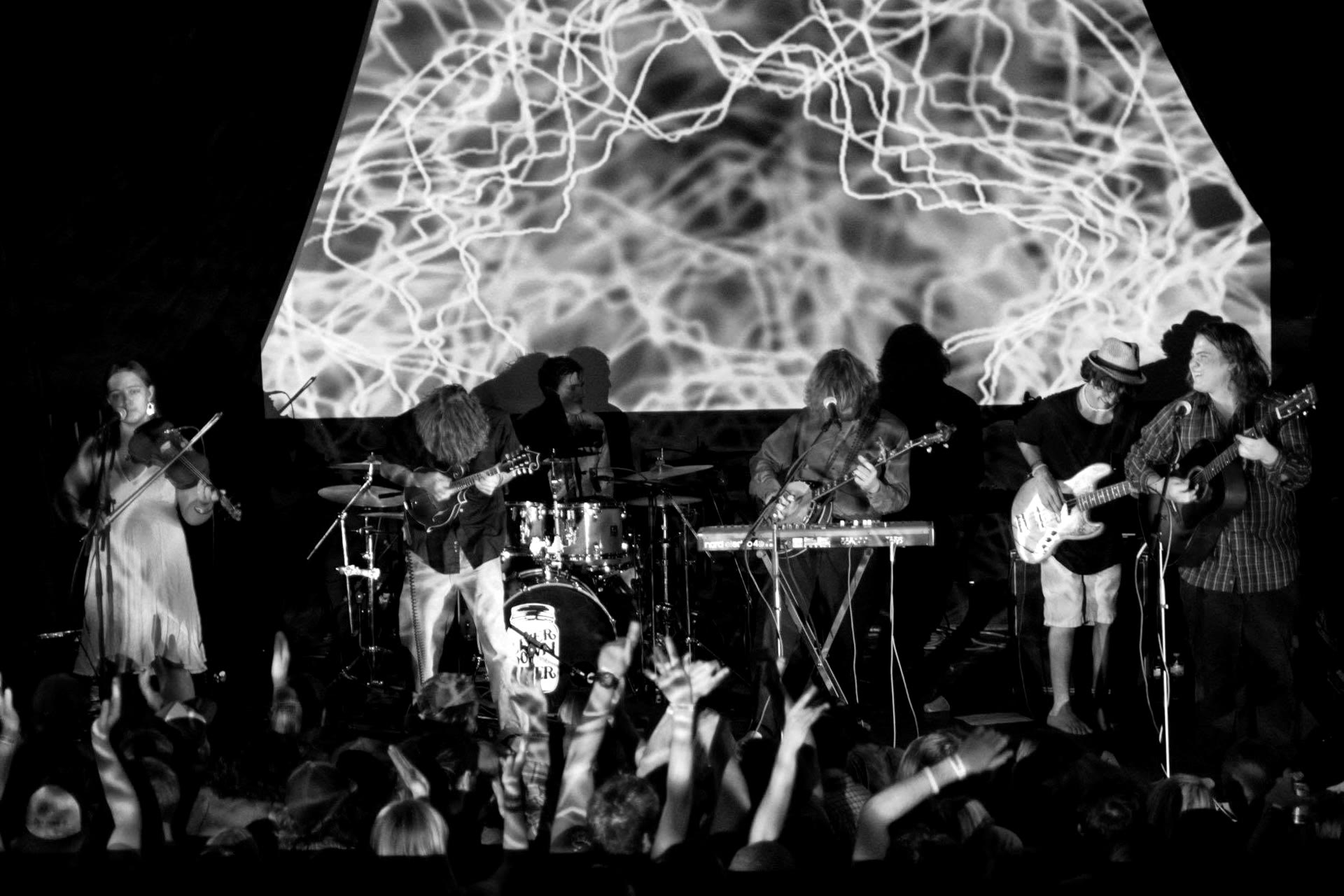 Full band under a purple lightning projection with crowd up front, black and white