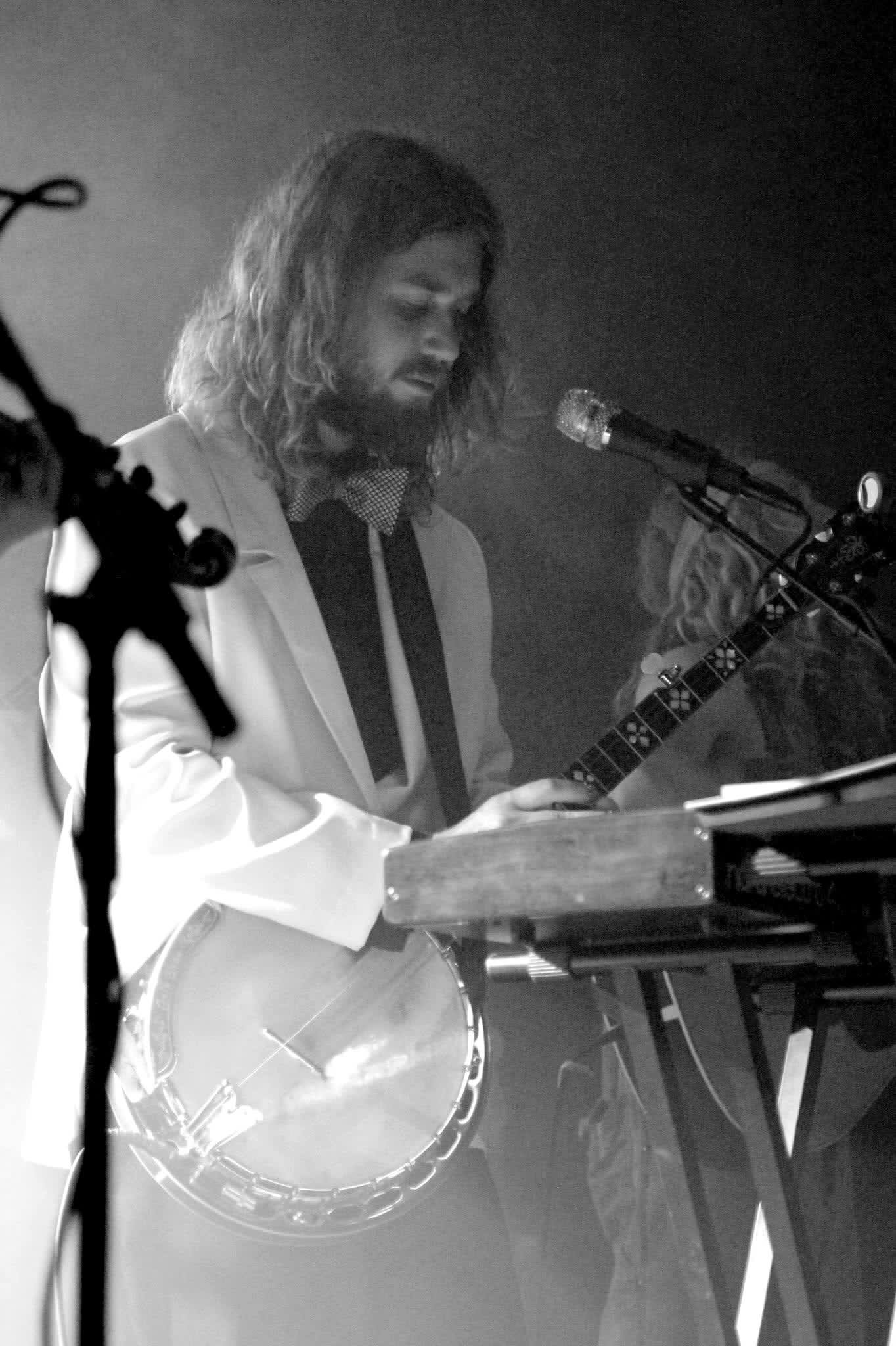 Tyler Rice in a white suit playing banjo over a red keyboard on a smoky stage