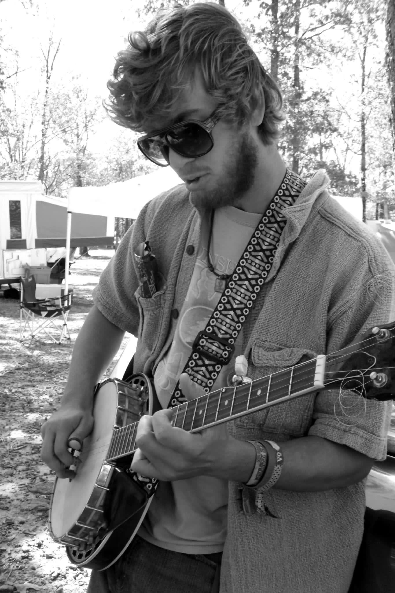 Tyler Rice with banjo and sunglasses at a wooded festival campground