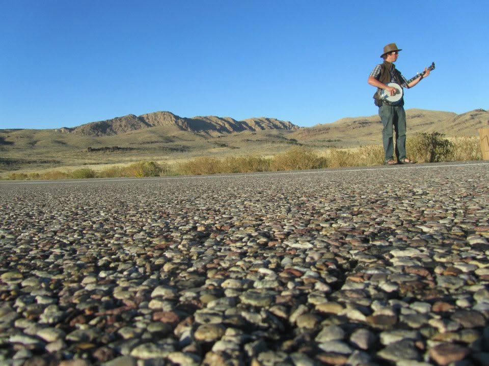 Color photo of Tyler Rice playing banjo alone on an empty desert highway