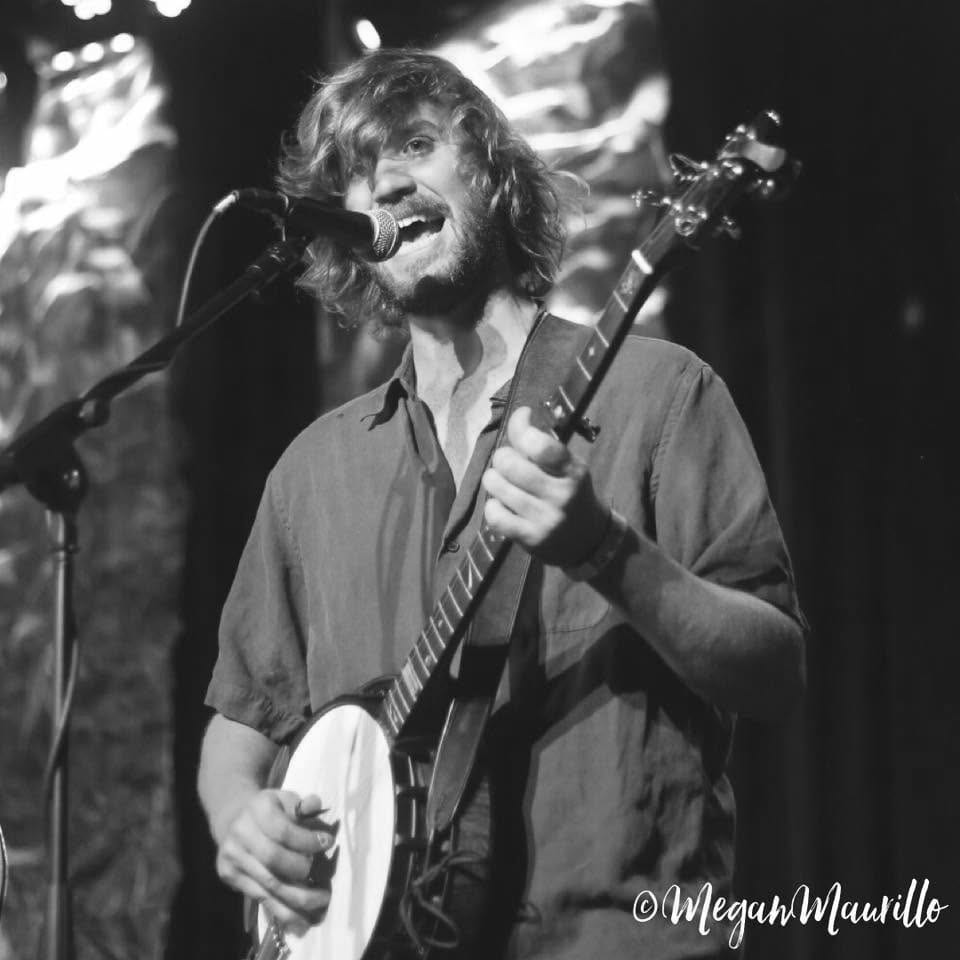 Tyler Rice singing hard into a microphone while playing banjo under green and gold stage lights